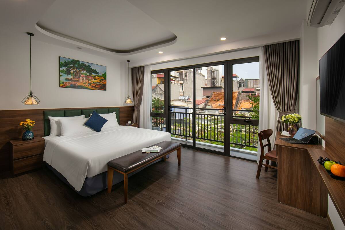 A modern hotel room with a large bed, a desk with a laptop, a view of the city through large windows, and a painting on the wall.