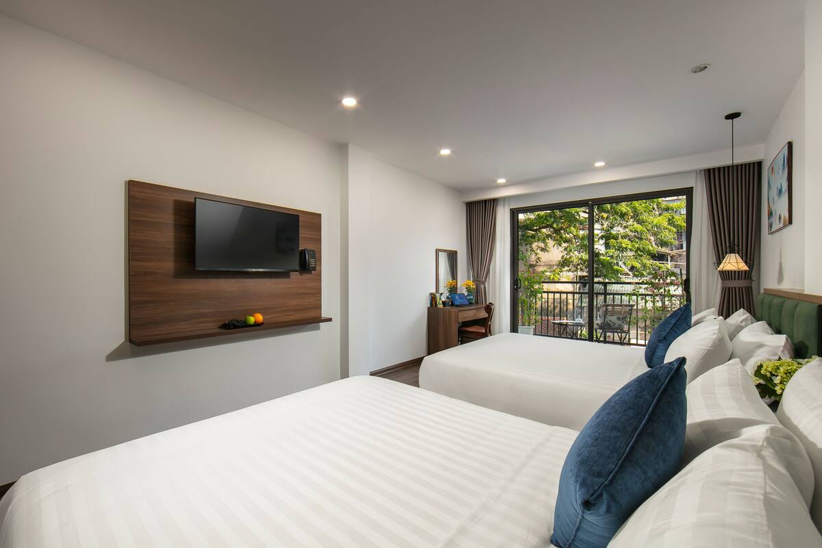 A hotel room with two beds, a TV mounted on the wall, a desk with a chair, and a balcony with a view of greenery.
