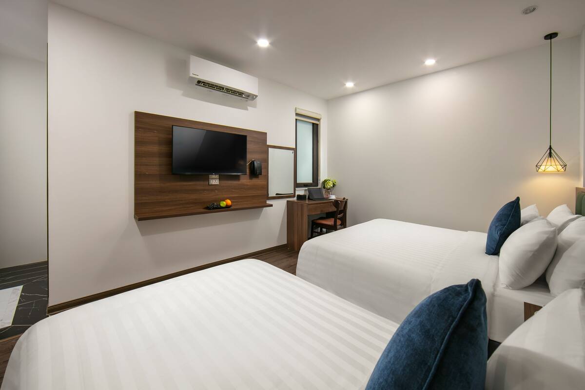 A hotel room with two beds, a TV mounted on the wall, a desk with a chair, and a hanging light fixture.