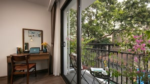 A room with a wooden desk, a laptop, a chair, and a view of a balcony with a railing and some plants.