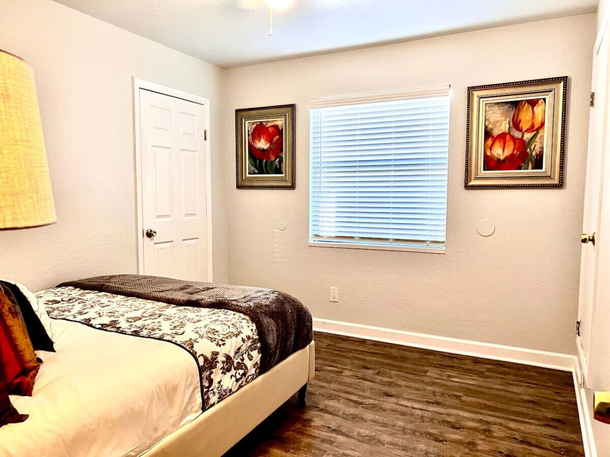 A bedroom with a bed, a door, a window with blinds, and two framed pictures on the wall.