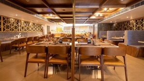 A modern dining area with wooden tables and chairs, a high ceiling with exposed beams, and a patterned wall design. - Hotel Park Elite (Gannavaram)