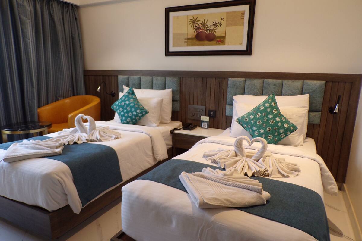 Een hotelkamer met twee bedden, elk voorzien van een blauwe dekbedovertrek, witte kussens en handdoeken gevouwen in de vorm van zwanen. Daarboven hangt een lijst met een afbeelding aan de muur, boven de bedden, met daaronder een klein tafeltje met een lamp en een stoel.