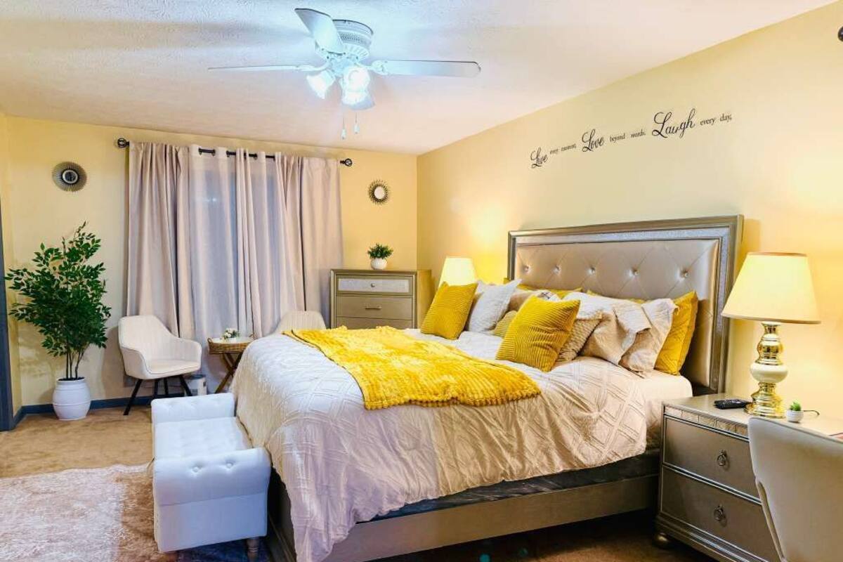 A bedroom with a bed, bedside tables, a chair, a desk, a ceiling fan, and a wall with a motivational quote.
