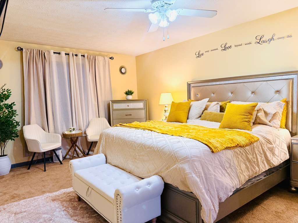 Een slaapkamer met een bed, nachtkastjes, een bankje, een plafondventilator, een kamerplant en een muur met motiverende tekst.
