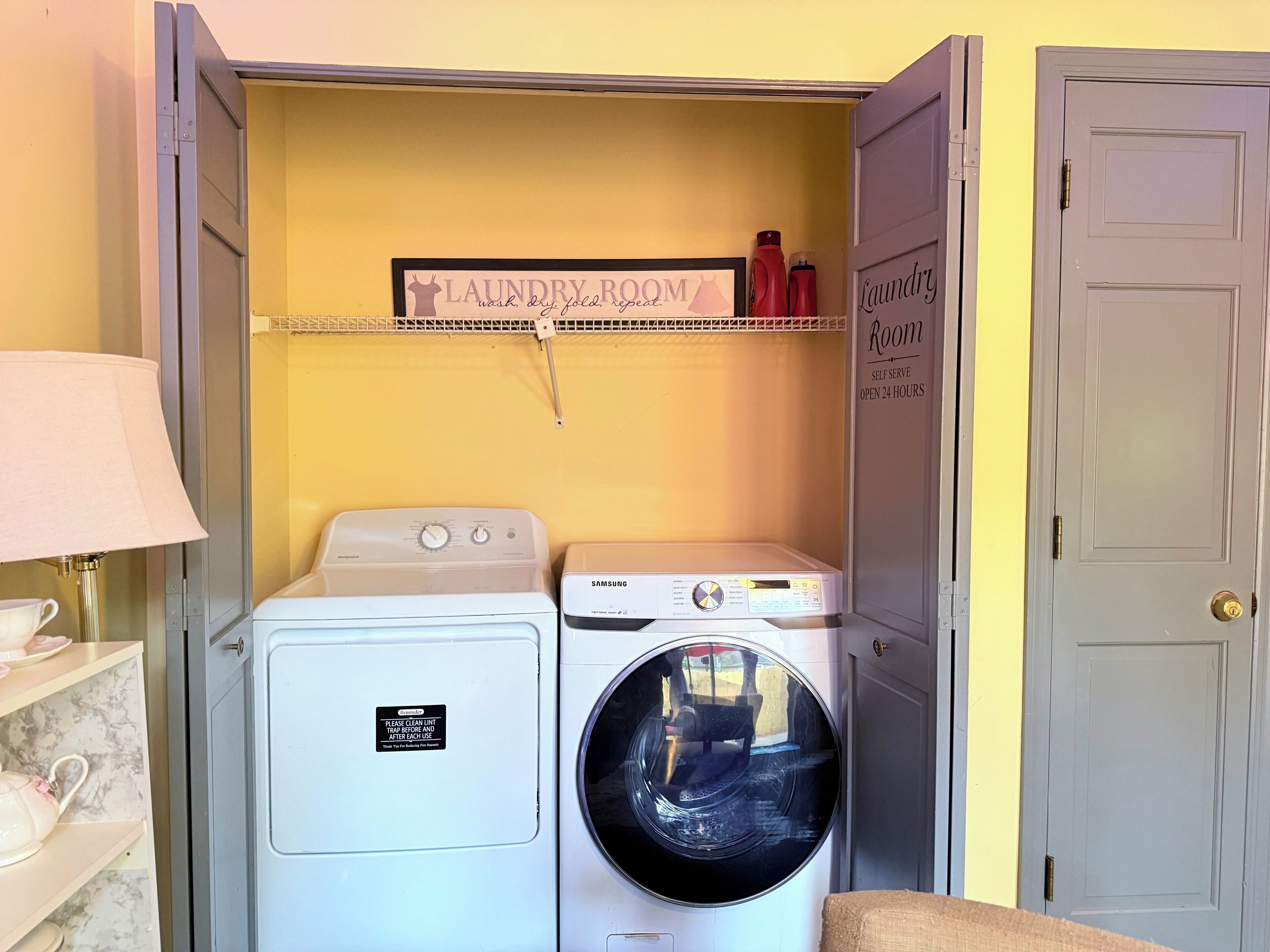 Une buanderie équipée de deux machines à laver et d’un sèche-linge, d’une étagère contenant des produits de lessive et d’un panneau indiquant qu’il s’agit d’un établissement en libre-service, ouvert 24 heures sur 24.