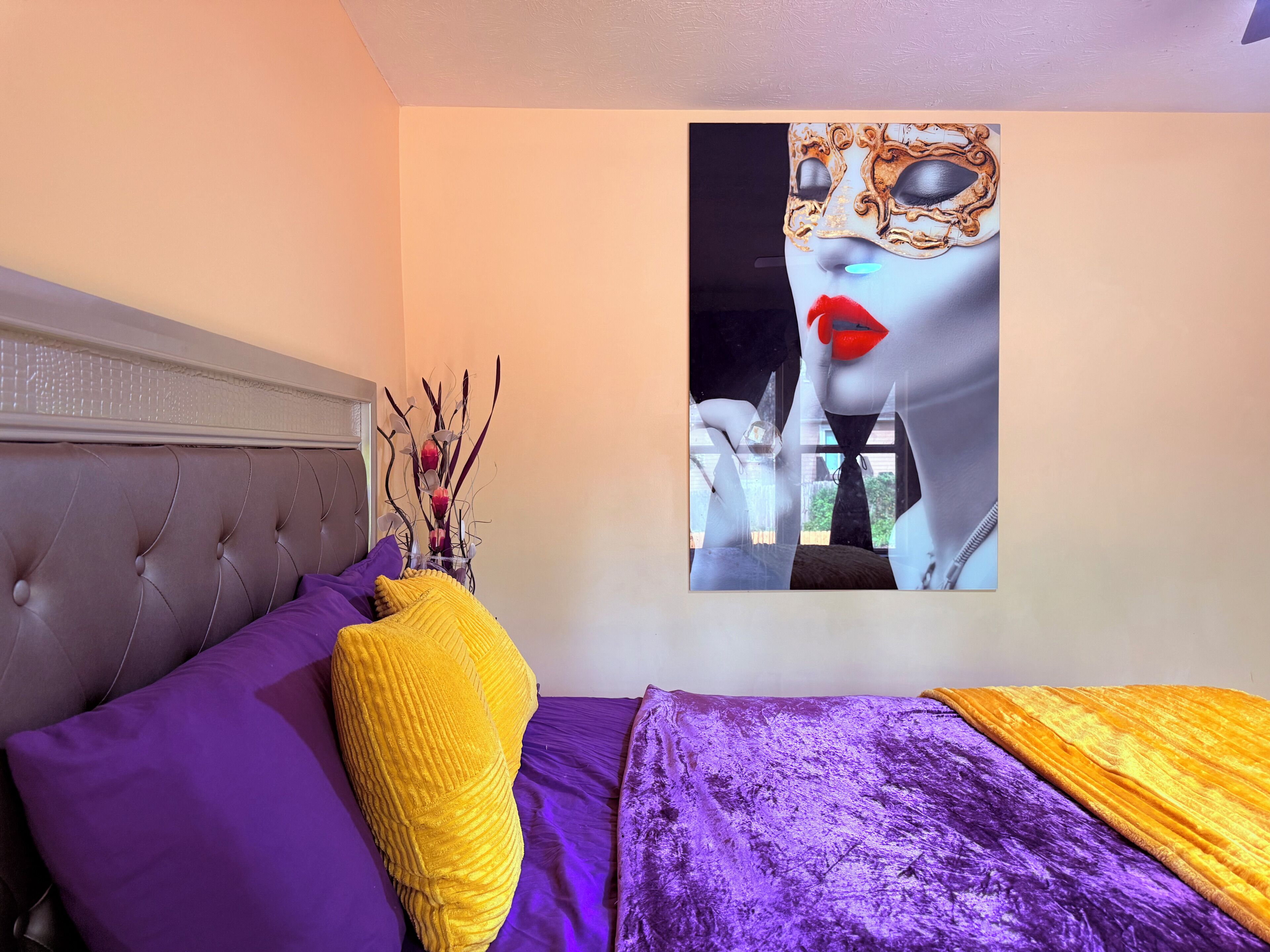 Un dormitori amb un capçal tapissat, roba de llit morada, un coixí decoratiu groc i una obra d'art penjada a la paret.