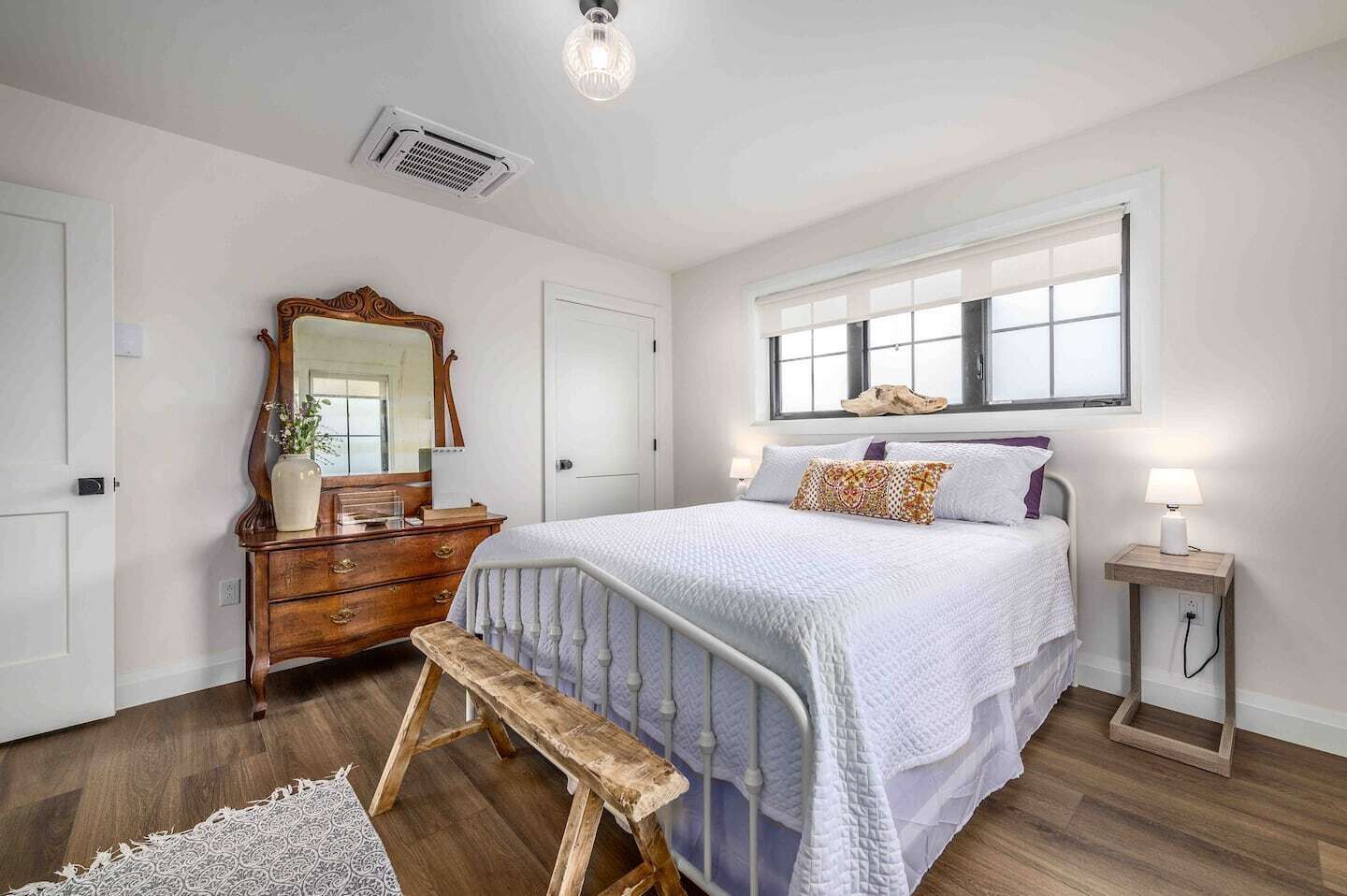 Une chambre à coucher comprenant un lit, une commode, un miroir, un banc, une table de chevet et une fenêtre.
