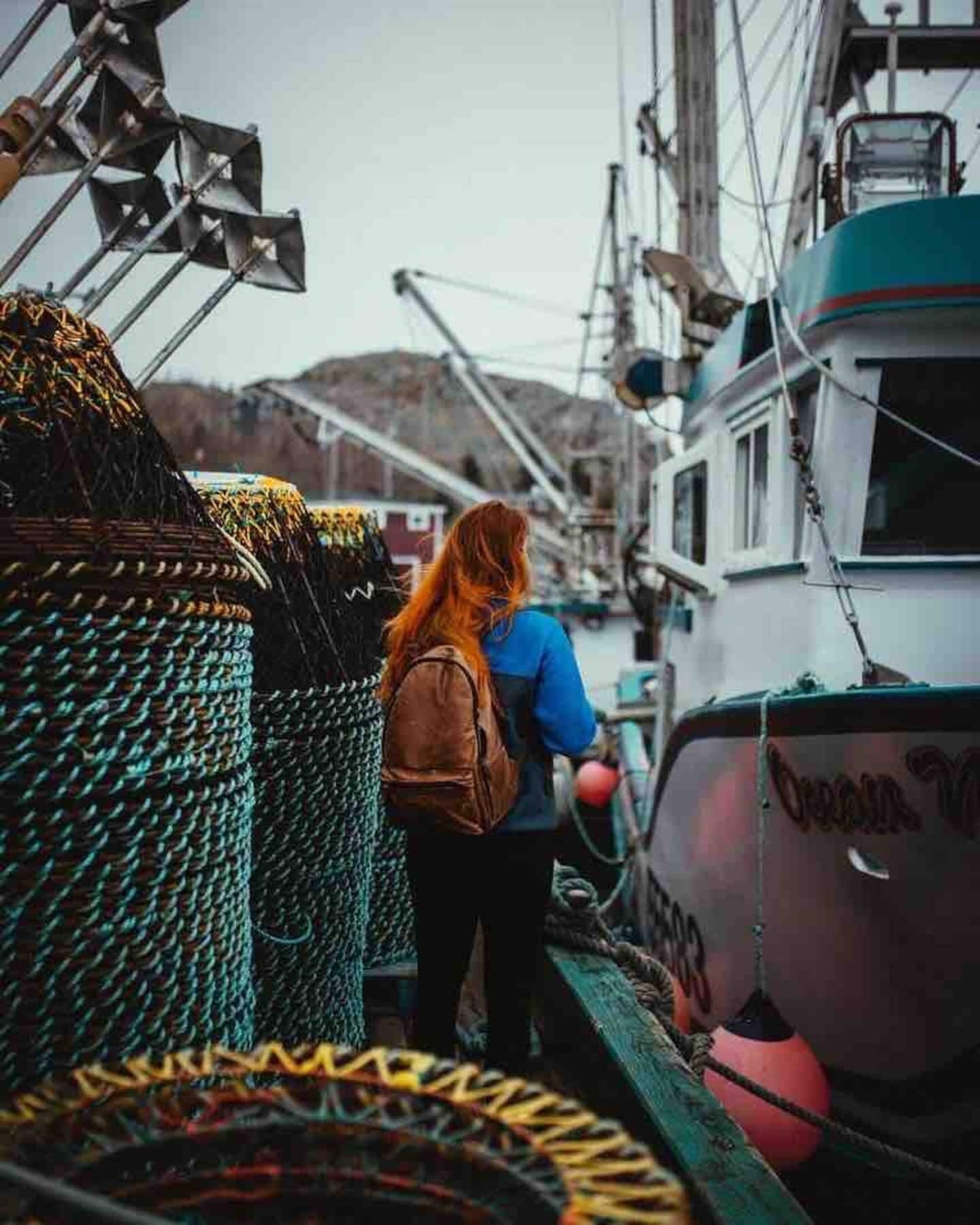Une personne se tient sur un quai, avec des filets de pêche, et un bateau est visible à l'arrière-plan.