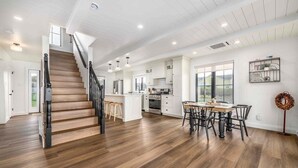 Una cocina amplia con armarios blancos, una zona de comedor con una mesa y sillas de madera, y una escalera con escalones de madera y barandillas negras.