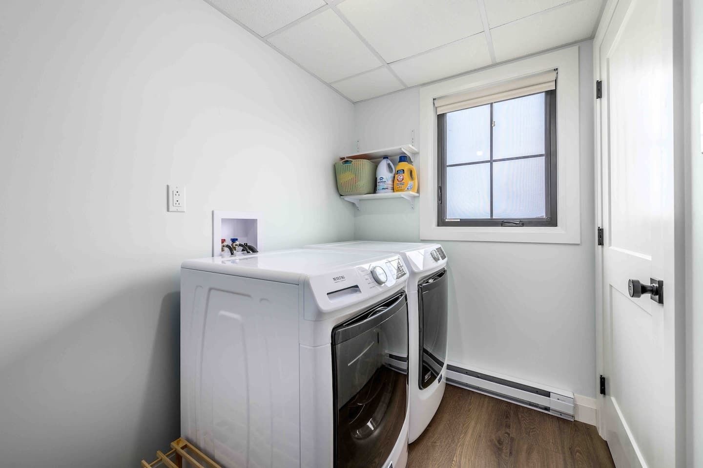 A laundry room with a stacked washer and dryer, a window, and a shelf with cleaning supplies.
