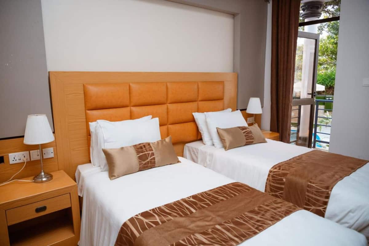 A hotel room with two beds, a wooden headboard, white bedding, a bedside lamp, and a view of the outdoors.