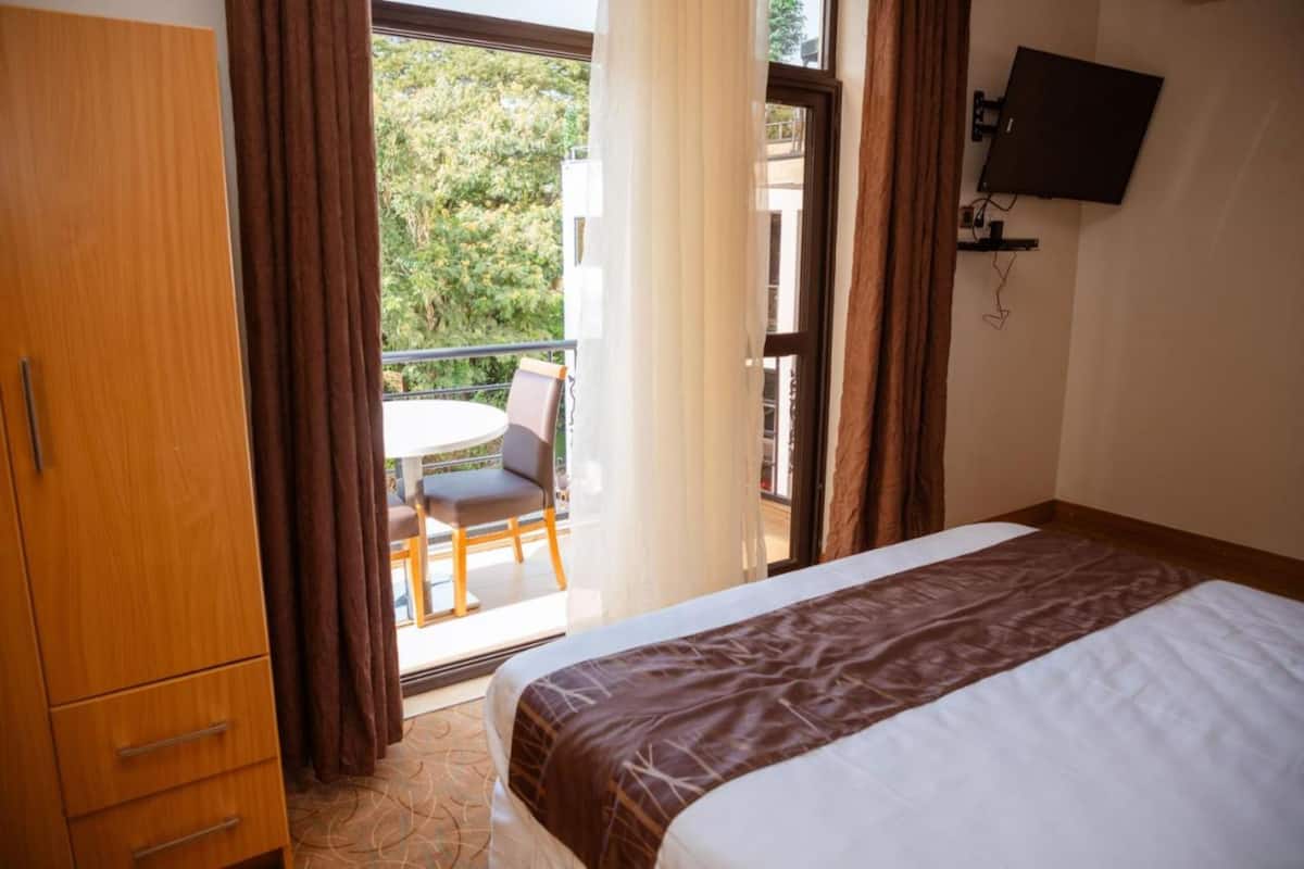 A hotel room with a bed, a wooden wardrobe, a balcony with a table and chairs, and a TV mounted on the wall.