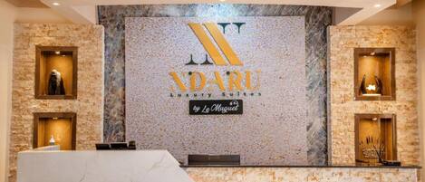 Un área de recepción con una pared de mármol, el logotipo de 'NADARU Luxury Suites by Le Muguet' y dos vitrinas con objetos.