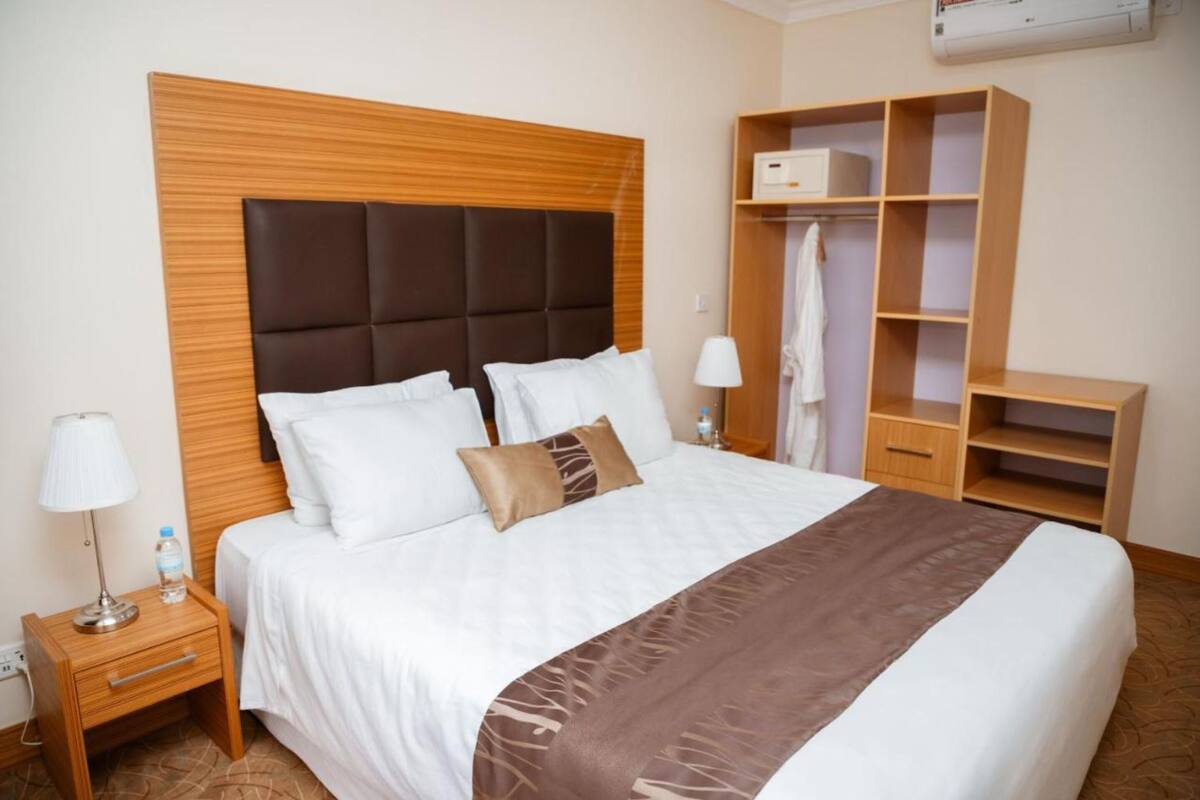 A hotel room with a large bed, a wooden headboard, a wardrobe, and a nightstand with a lamp and a bottle of water.