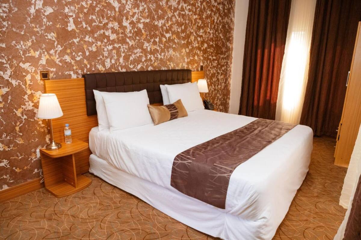 A hotel room with a large bed, two bedside tables with lamps, a wooden headboard, and a patterned carpet.
