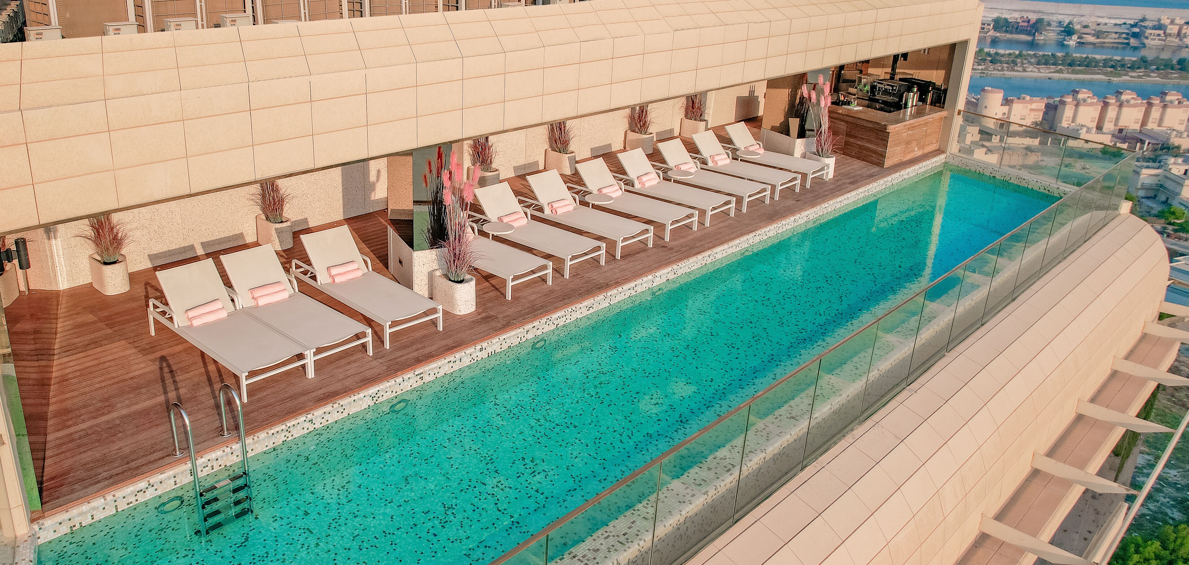 Piscina infinita en la azotea con sillones de descanso y plantas en macetas, con vista a un paisaje urbano.