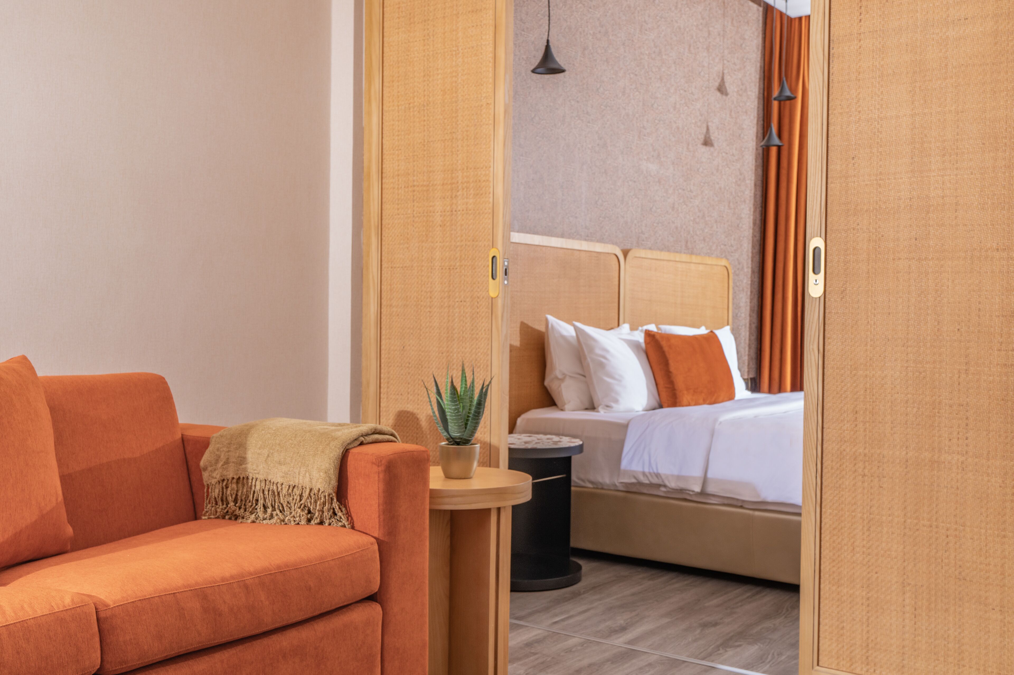 A modern hotel room with a sofa, a small round table with a plant, a bed with white bedding and orange pillows, and a wooden wardrobe.