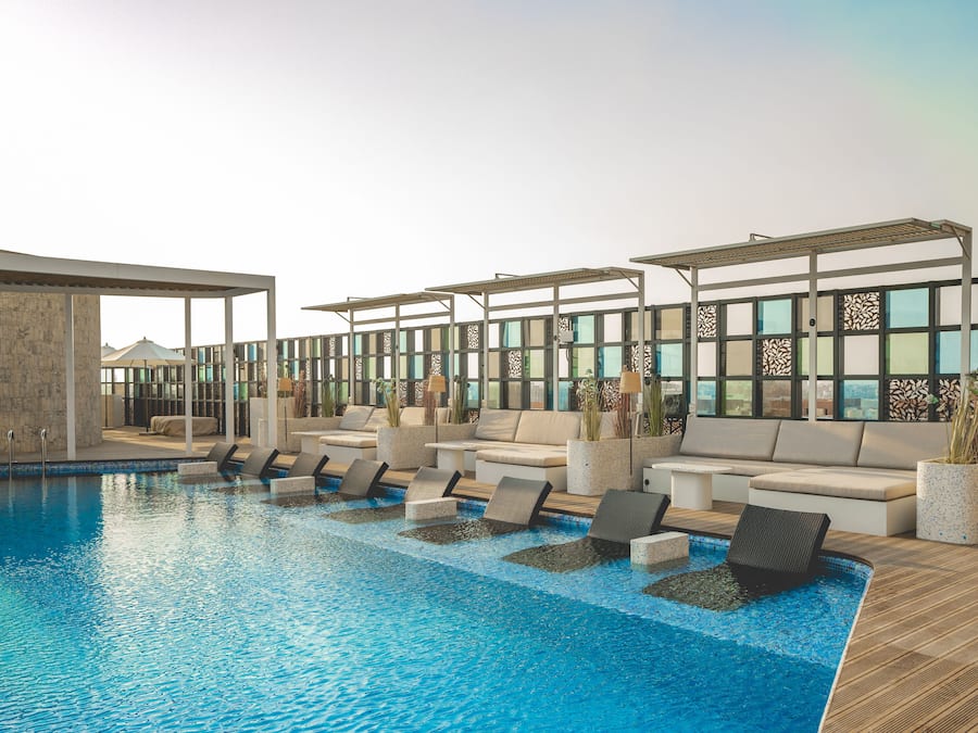 A rooftop pool area with lounge chairs and seating, surrounded by large windows and decorative panels.