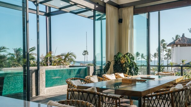 Un espacio interior moderno con pared de vidrio que da a una piscina y palmeras.
