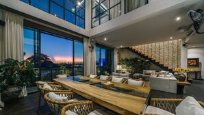 Amplia sala de estar con una larga mesa de madera, sillas de mimbre y un sofá. Se observa una escalera con pared de piedra, y grandes ventanales que ofrecen vistas al atardecer.