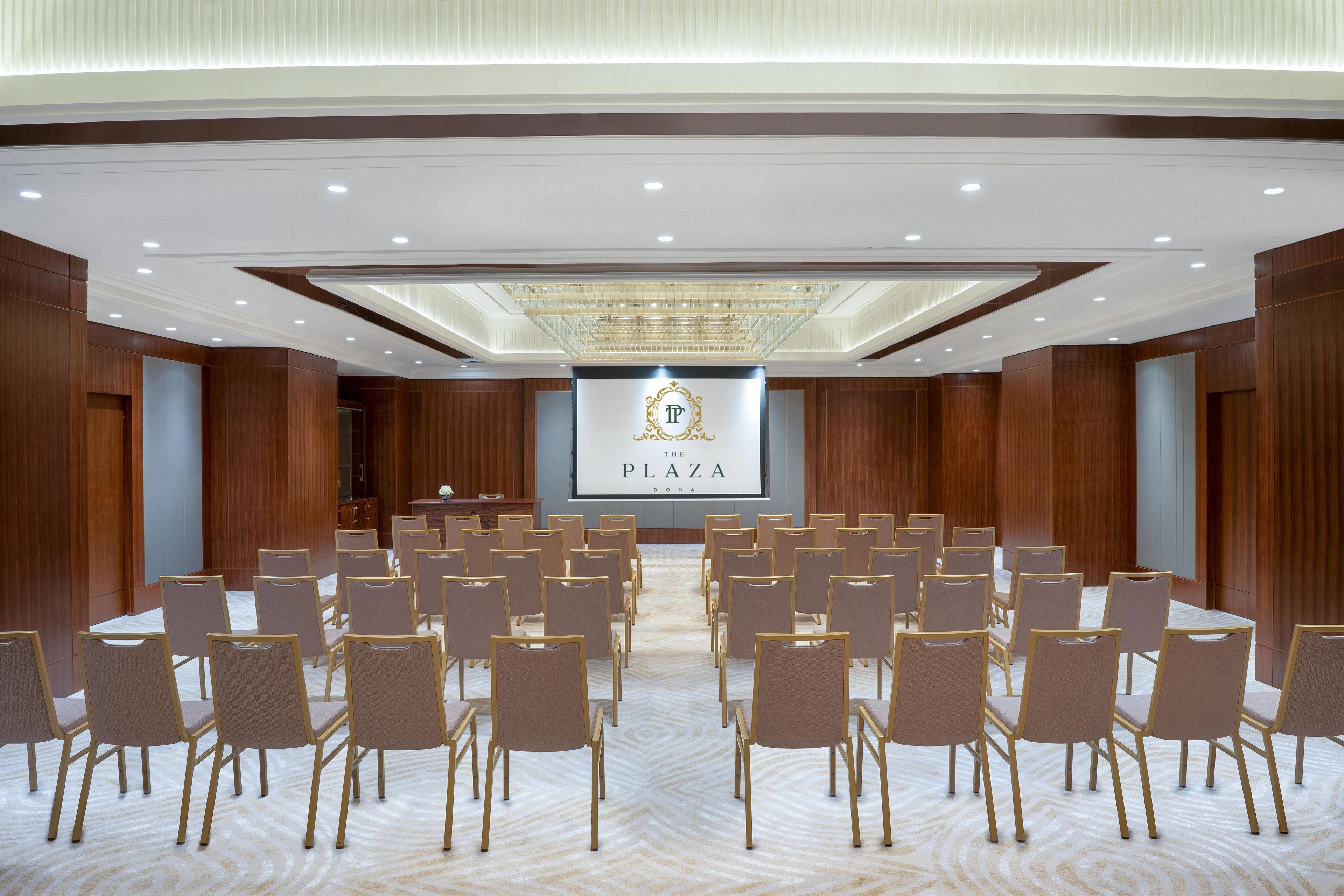 Una sala conferenze con file di sedie rivolte verso un podio con uno schermo che mostra 'THE PLAZA DOHA'.