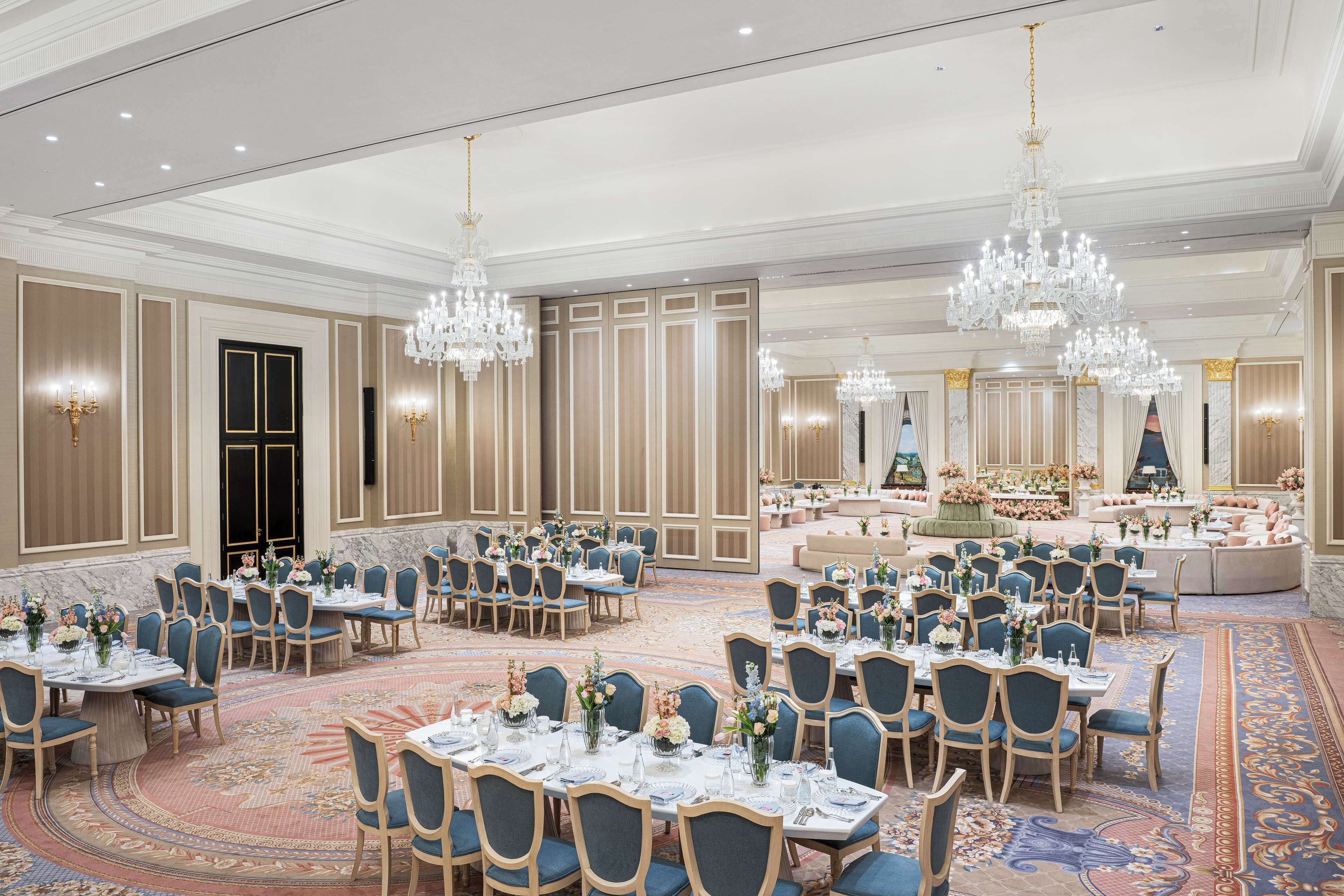 A spacious banquet hall with multiple round tables, blue upholstered chairs, and ornate chandeliers.