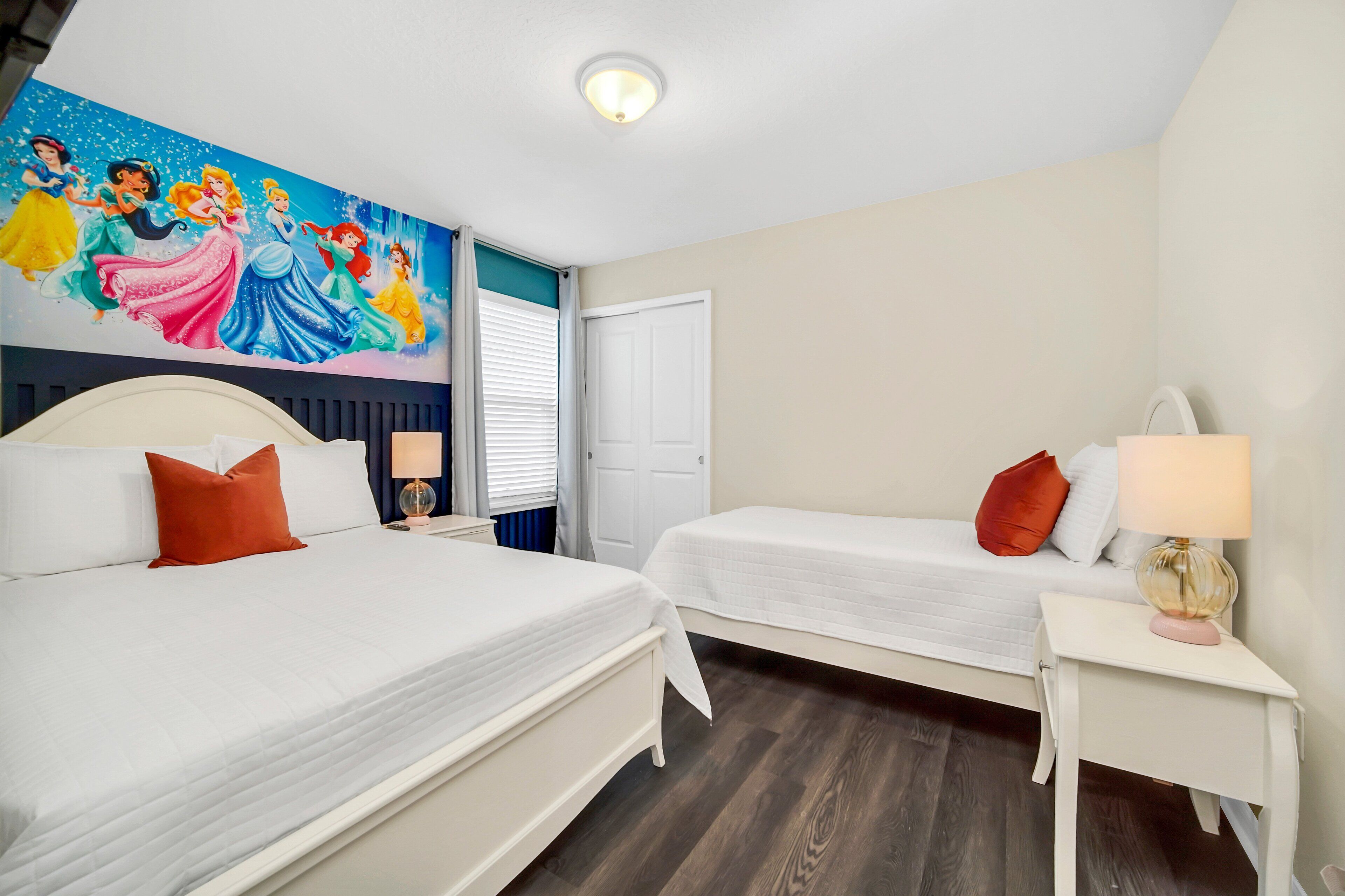 Habitación de hotel con dos camas, un mural de princesas Disney, una puerta blanca y una mesita de noche con lámpara.