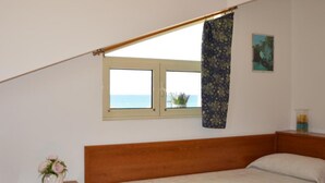 Une chambre avec un lit en bois, une table de chevet ornée d’un vase de fleurs, une fenêtre donnant sur la mer et un tableau accroché au mur.