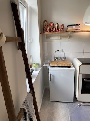 A compact kitchen with a white refrigerator, a small oven, and a wooden cutting board on top. There are decorative items on a shelf above the oven, and a window with a curtain partially visible. - Good times in St. Goar (Sankt Goar)