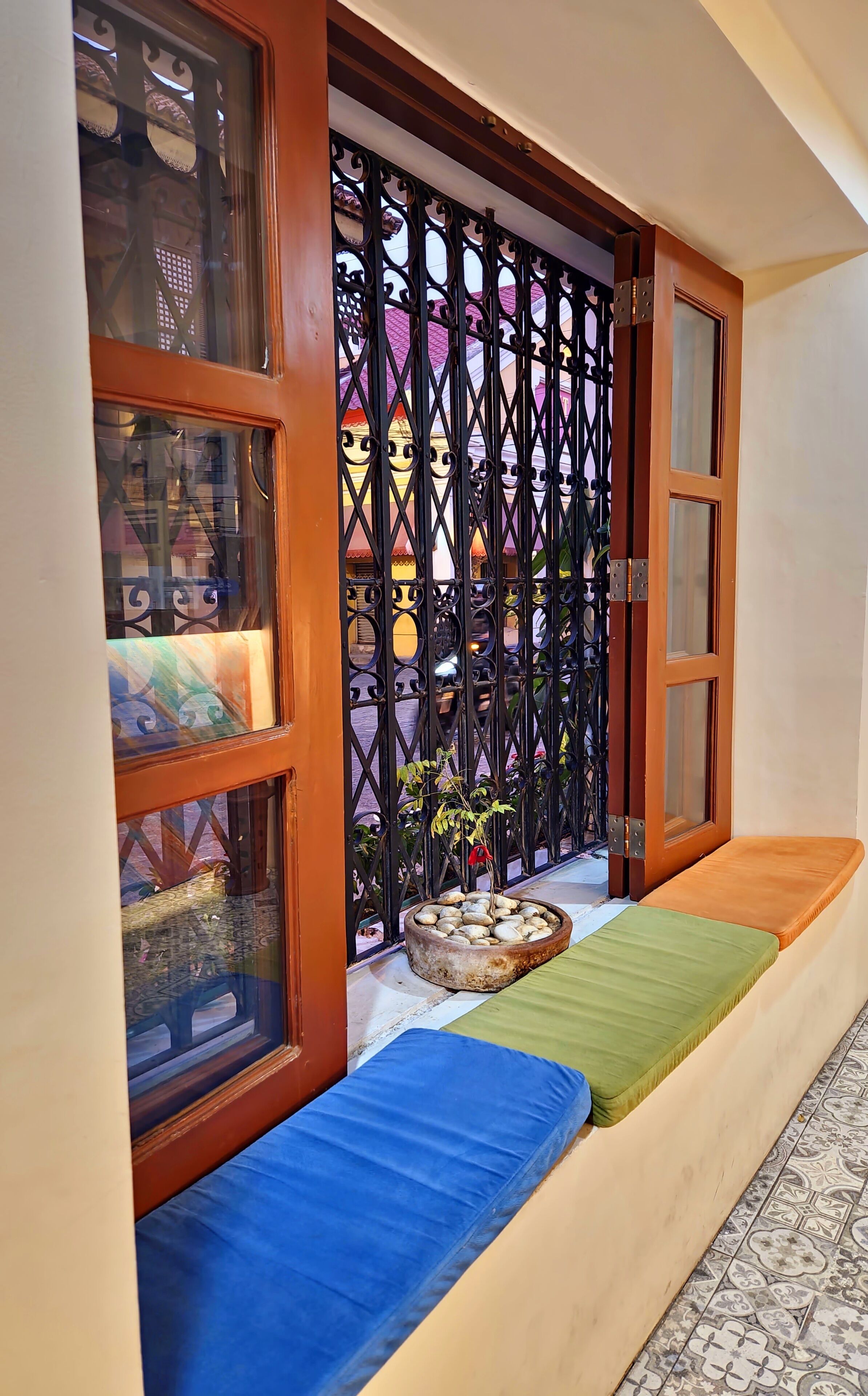 A room with a wooden door, a window with a decorative grill, a bench with cushions, and a tiled floor.