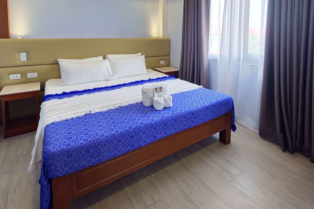 A neatly made bed with a blue and white bedspread, two white pillows, and a wooden headboard. There is a wooden nightstand with a lamp beside the bed. The room has light-colored wooden flooring and a window with curtains.