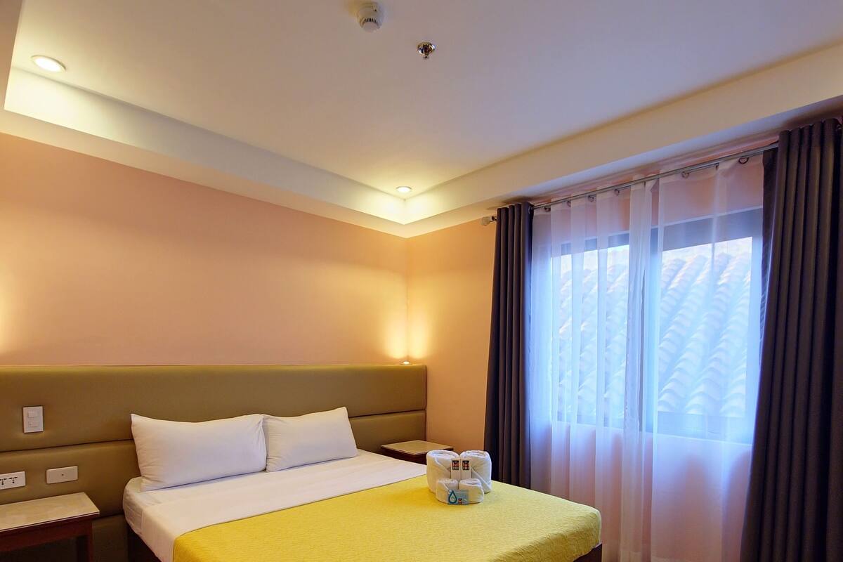 A hotel room with a large bed, a bedside table, a nightstand, a window with curtains, and a ceiling light.