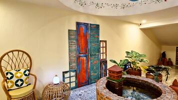 Un interior amplio con una escalera de madera curva, una chimenea decorativa, baldosas de suelo con motivos y una silla de mimbre con cojín estampado.