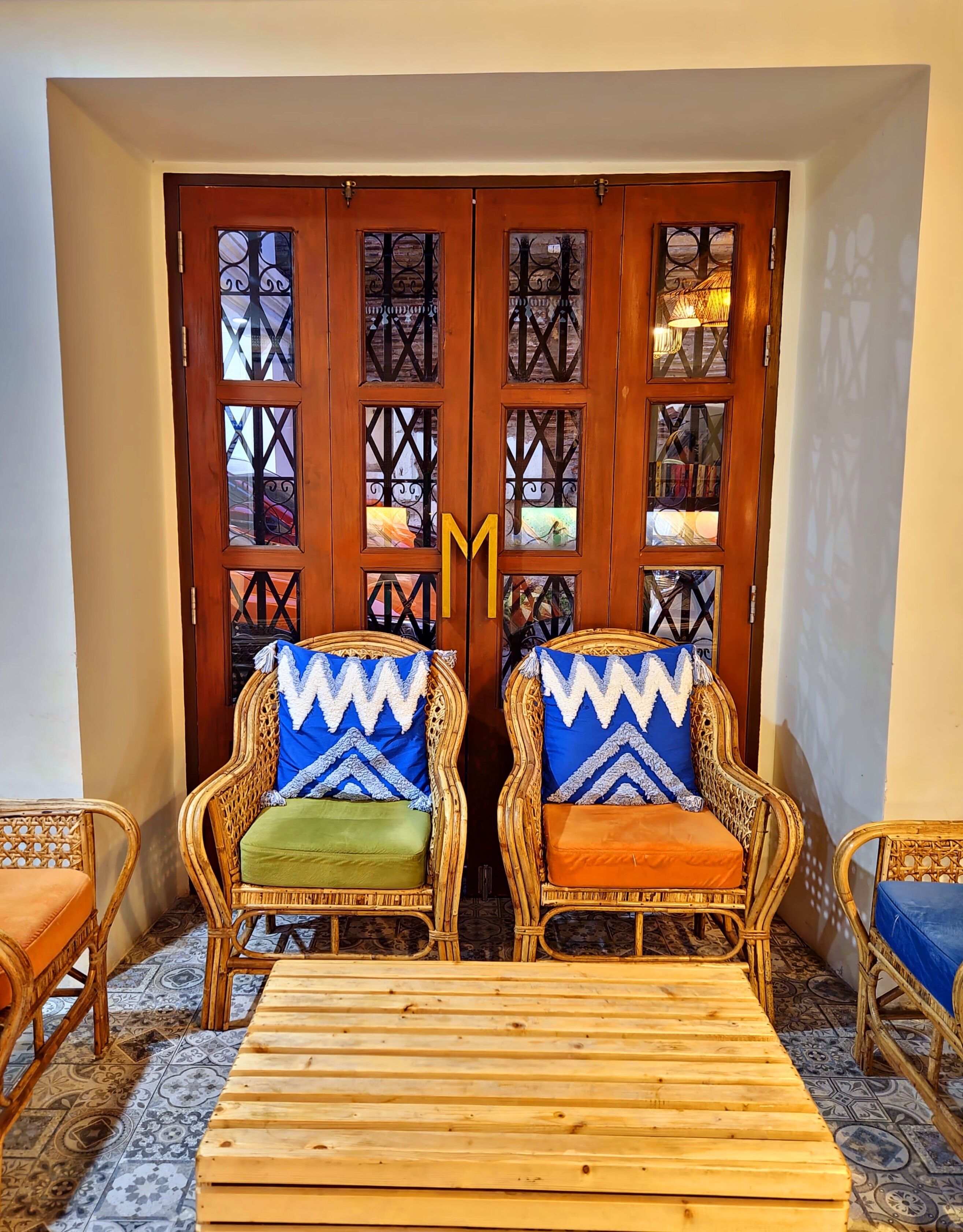 Una habitación con dos sillones de mimbre con cojines estampados, una mesa de madera y una gran puerta de madera con paneles de vidrio decorativos.