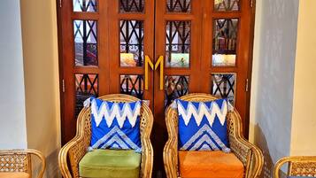 Una habitación con dos sillones de mimbre con cojines estampados, una mesa de madera y una gran puerta de madera con paneles de vidrio decorativos.