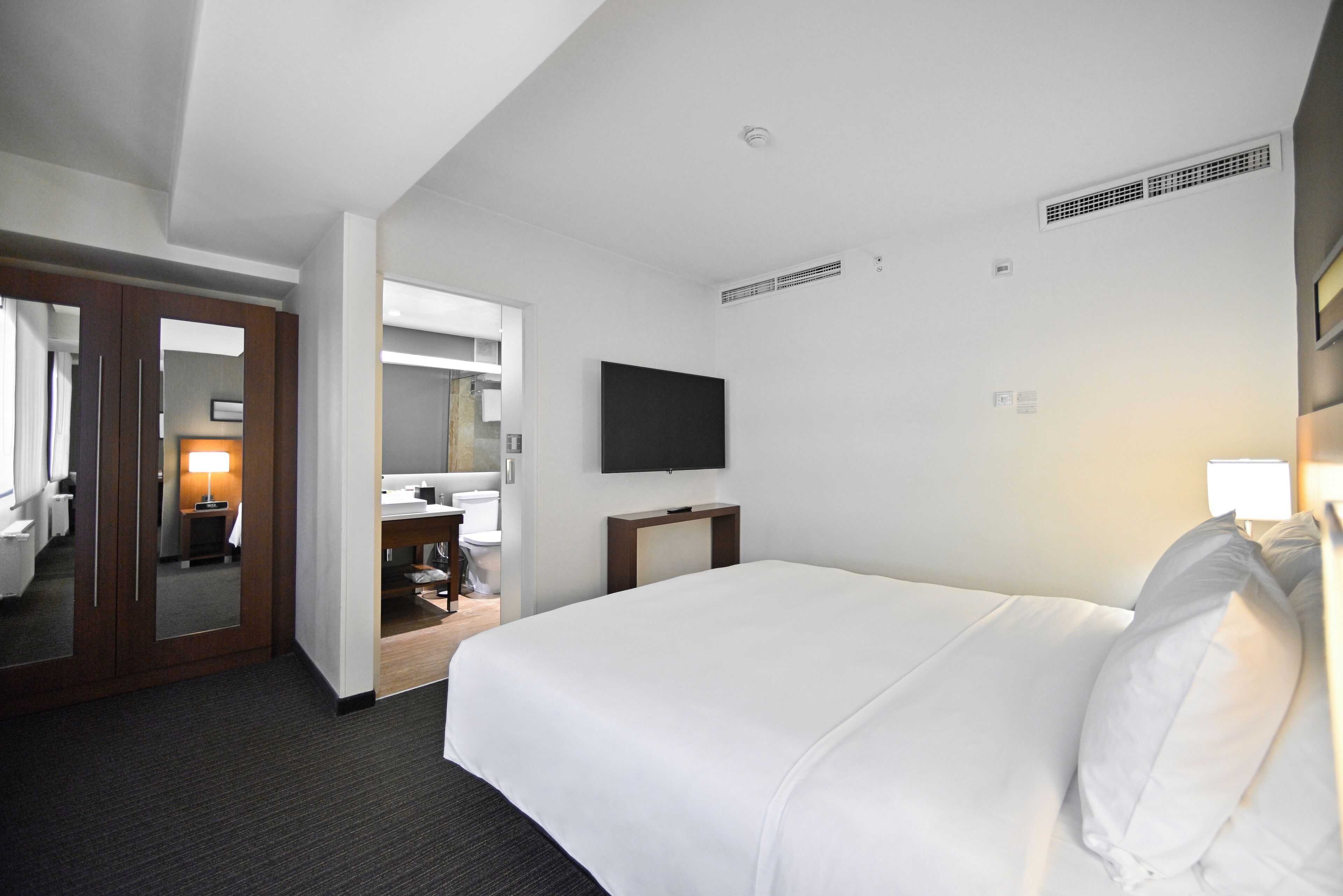 Une chambre d’hôtel avec un grand lit, un téléviseur à écran plat, un miroir et une salle de bain visible à travers une porte ouverte.