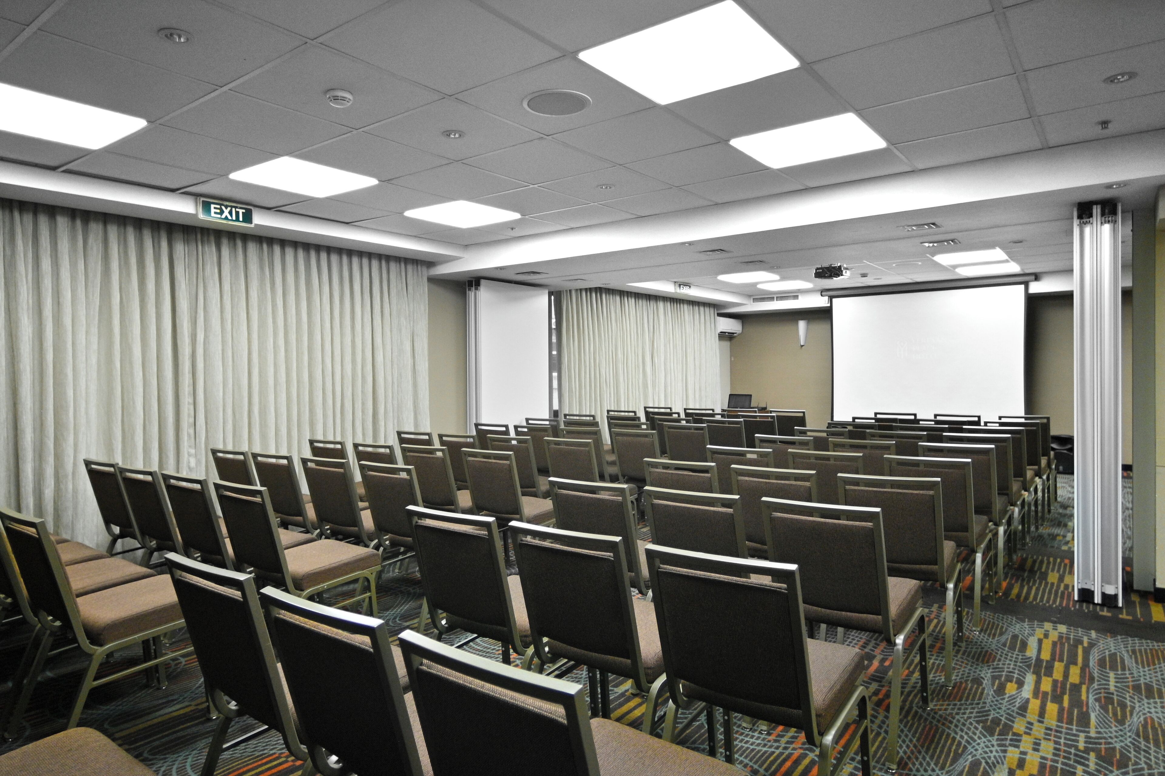 A conference room with rows of chairs, an exit sign, a projector screen, and a carpeted floor.