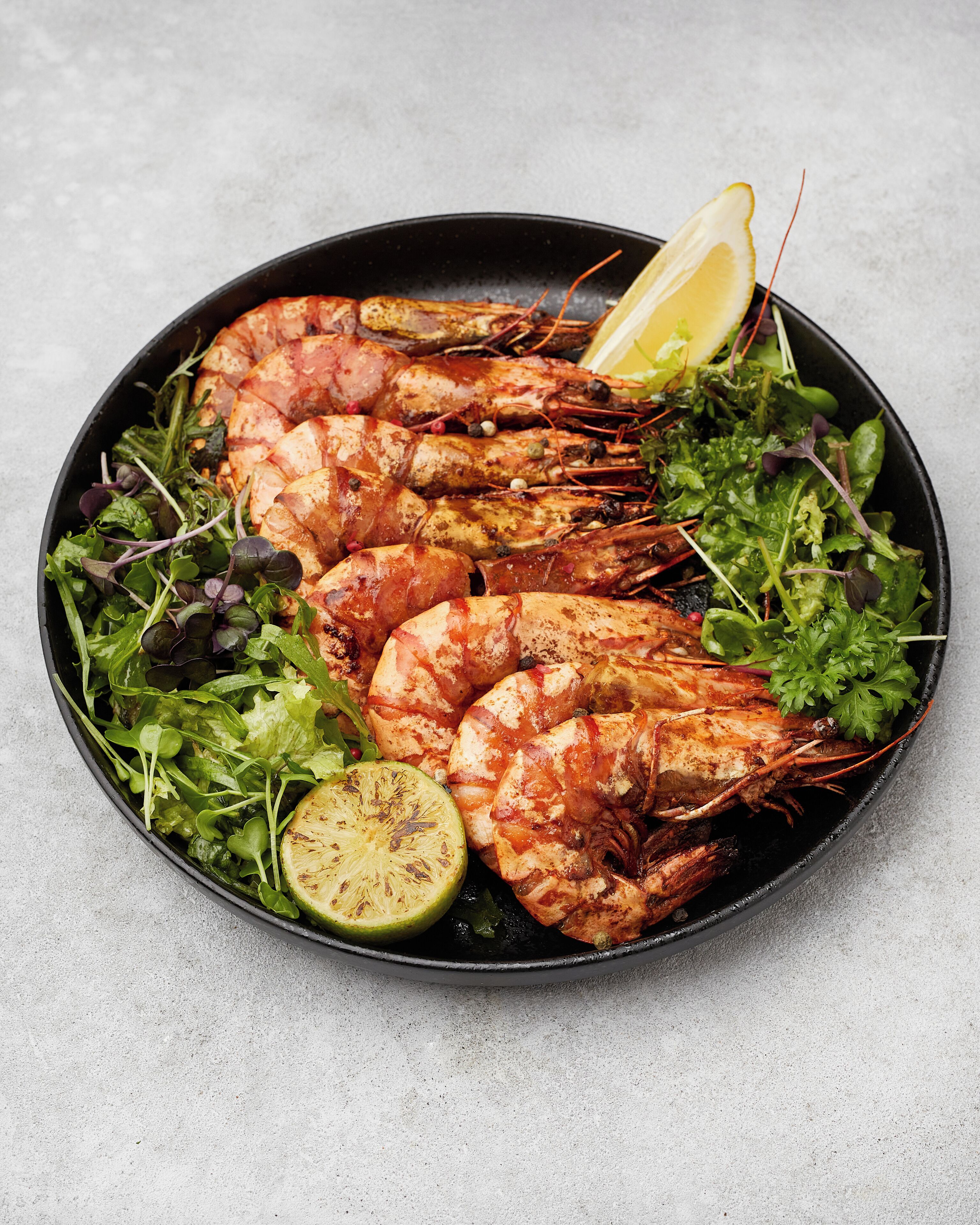 A bowl of cooked shrimp with greens and a slice of lime.
