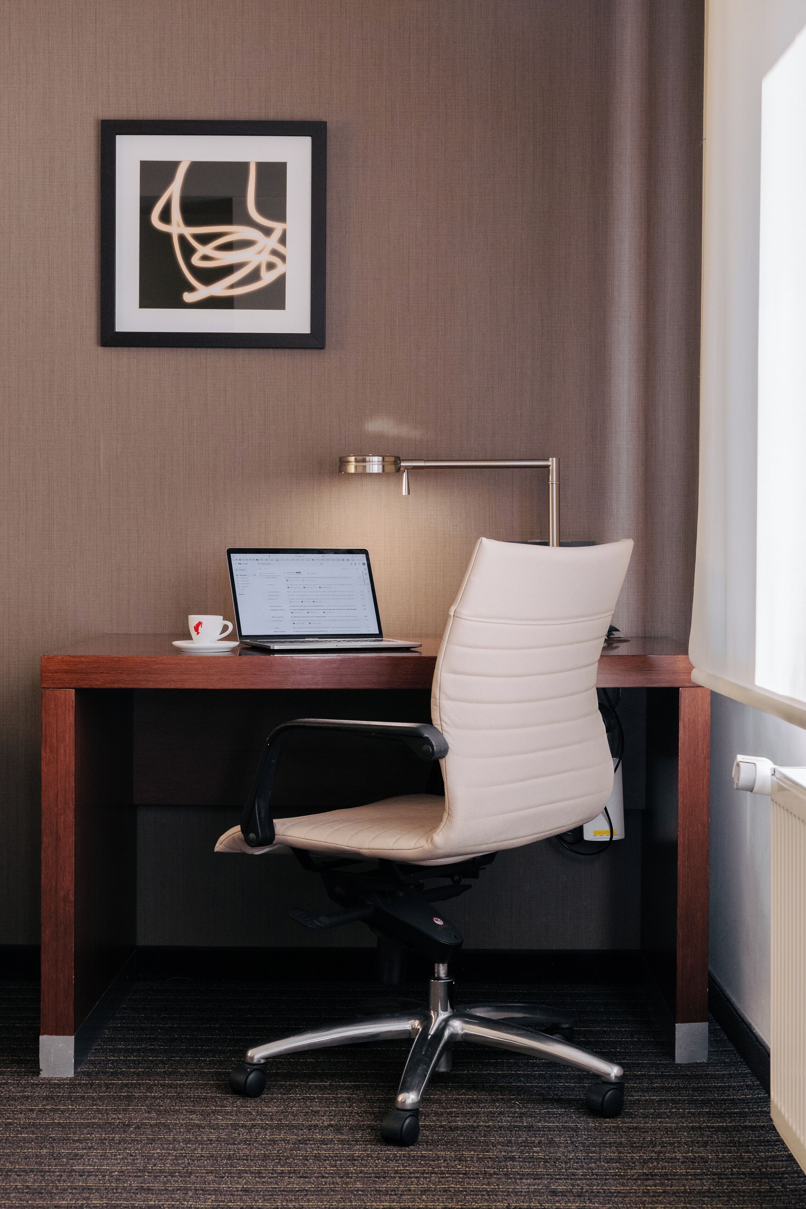 Une chambre d’hôtel avec un bureau en bois, un ordinateur portable, une tasse blanche, une chaise moderne et une œuvre d’art abstraite encadrée accrochée au mur.