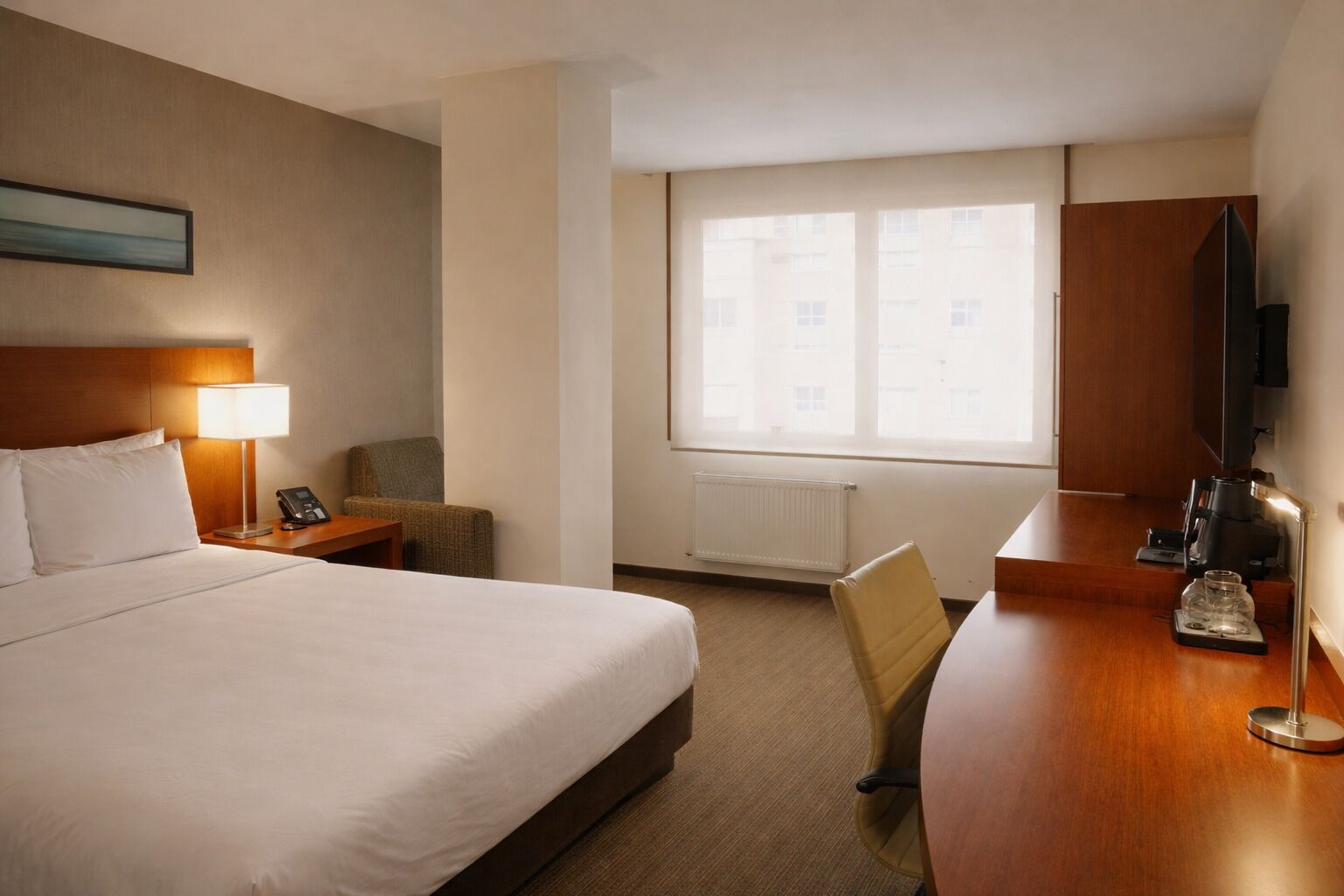 A hotel room with a bed, bedside lamp, chair, desk with coffee maker, TV, and a window with a view of buildings.