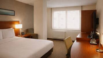 Quarto de hotel com cama, luminária de cabeceira, poltrona, escrivaninha com cafeteira, televisão e janela com vista para edifícios.