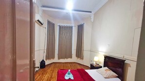 Um quarto com piso de madeira, uma cama com cobertor vermelho, uma mesa de cabeceira com abajur e uma janela com cortinas.