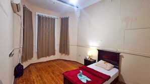Um quarto com piso de madeira, uma cama com cobertor vermelho, uma mesinha de cabeceira com abajur e uma janela com cortinas.