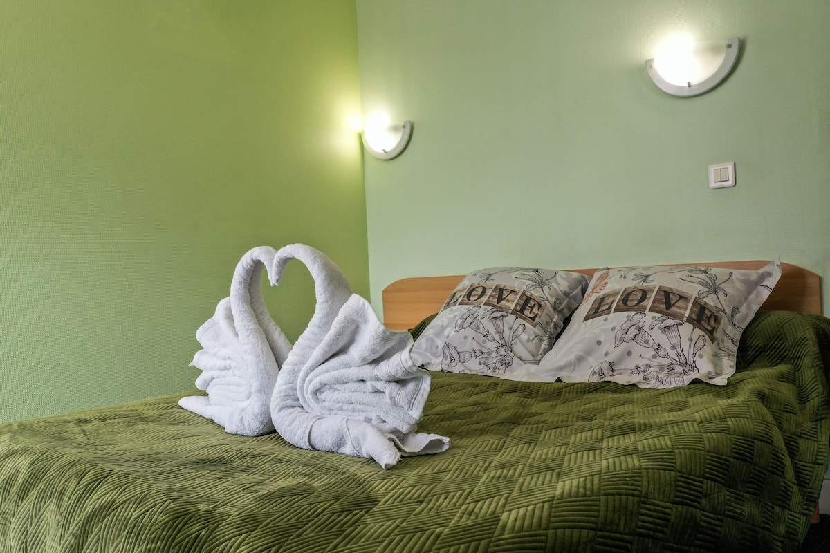 Ein Zimmer mit grüner Wand, einem Bett mit grüner Steude, zwei Kissen mit Blumenmuster und einem zu Schwänen geformten Handtuch.
