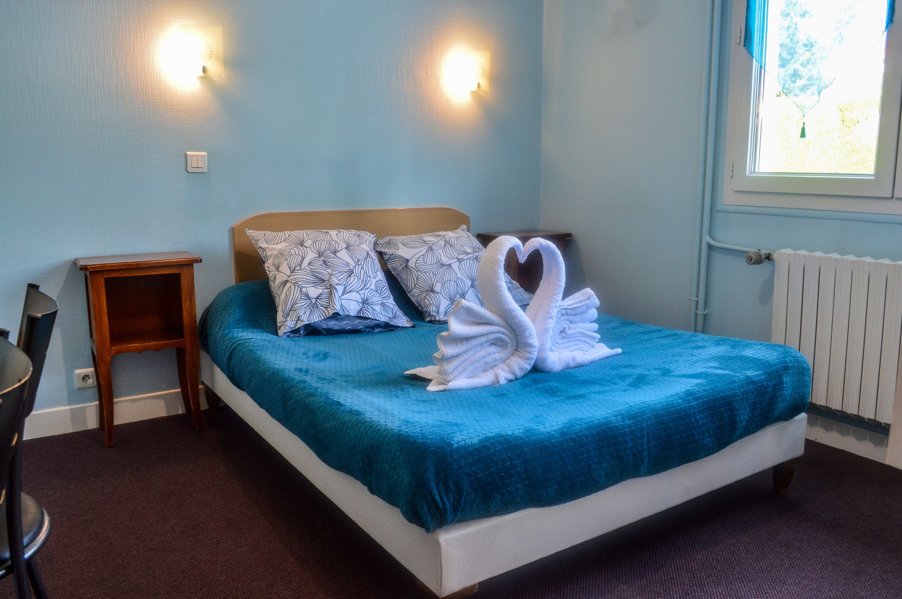 A neatly made bed with a blue bedspread and two patterned pillows, a small wooden nightstand, and a wall-mounted light fixture.