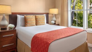 Quarto de hotel com uma cama grande, duas mesas de cabeceira, luminárias, uma janela com vista para vegetação e um cobertor vermelho.