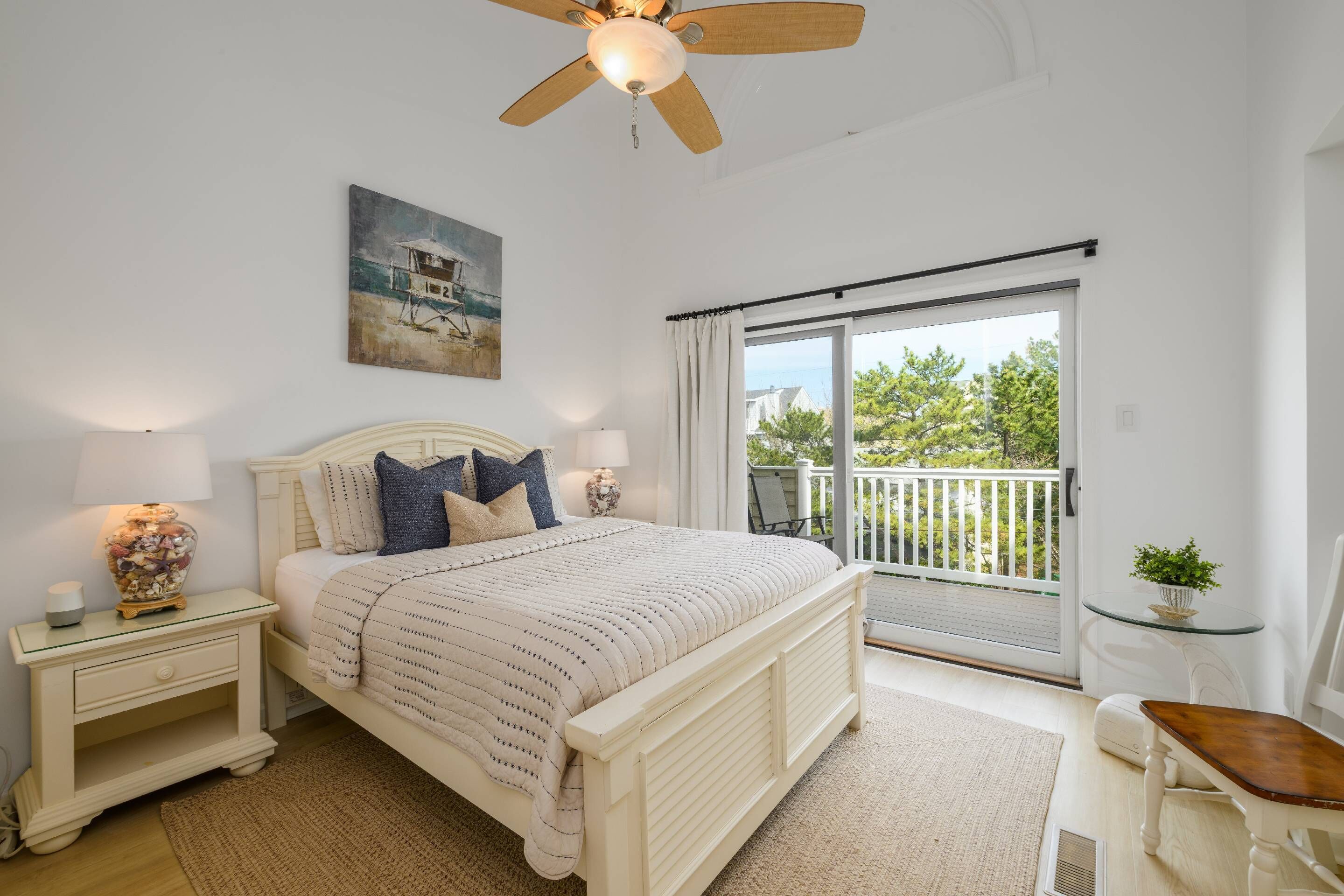 A bedroom with a bed, nightstands, a ceiling fan, a painting, a balcony with a view, and a small table with a plant.