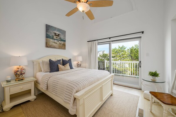 A bedroom with a bed, nightstands, a ceiling fan, a painting, a balcony with a view, and a small table with a plant.