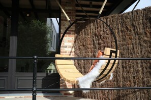 A wooden swing chair with a white feathered tail, attached to a rope, against a backdrop of a textured wall and a metal railing.